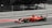 A red Formula 1 race car speeds along a track. The car is adorned with various logos and sponsors, featuring sleek aerodynamic design elements. The background is a blurred mix of gray and black, emphasizing the motion and speed of the vehicle.