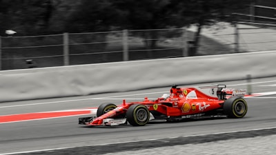 A red Formula 1 race car speeds along a track. The car is adorned with various logos and sponsors, featuring sleek aerodynamic design elements. The background is a blurred mix of gray and black, emphasizing the motion and speed of the vehicle.
