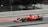 A red Formula 1 race car speeds along a track. The car is adorned with various logos and sponsors, featuring sleek aerodynamic design elements. The background is a blurred mix of gray and black, emphasizing the motion and speed of the vehicle.