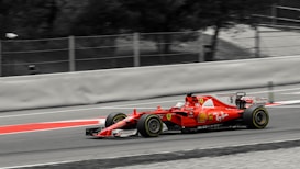 A red Formula 1 race car speeds along a track. The car is adorned with various logos and sponsors, featuring sleek aerodynamic design elements. The background is a blurred mix of gray and black, emphasizing the motion and speed of the vehicle.