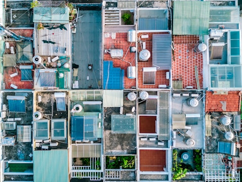 Aerial view of several rooftops with various structures like water tanks, solar panels, and different colored tiles. Some roofs have greenery, and others display antennas and vents. The rooftops are bordered by narrow pathways.