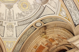 An intricately decorated ceiling features detailed artwork and patterns. The design includes ornate carvings, geometric shapes, and classical figures. The color palette comprises gold, beige, and black accents, lending a luxurious and historic feel.