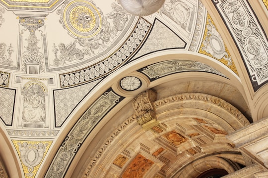 An intricately decorated ceiling features detailed artwork and patterns. The design includes ornate carvings, geometric shapes, and classical figures. The color palette comprises gold, beige, and black accents, lending a luxurious and historic feel.