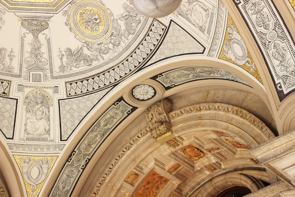 An intricately decorated ceiling features detailed artwork and patterns. The design includes ornate carvings, geometric shapes, and classical figures. The color palette comprises gold, beige, and black accents, lending a luxurious and historic feel.