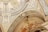 An intricately decorated ceiling features detailed artwork and patterns. The design includes ornate carvings, geometric shapes, and classical figures. The color palette comprises gold, beige, and black accents, lending a luxurious and historic feel.