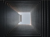 A geometric view looking upward through the center of a multi-story building. The image focuses on the square shape formed by the inner walls, leading to an open sky at the top. The architectural details create a repetitive pattern, enhancing the sense of depth.