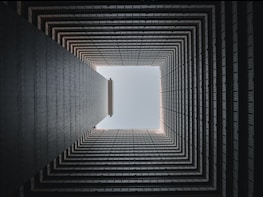 A geometric view looking upward through the center of a multi-story building. The image focuses on the square shape formed by the inner walls, leading to an open sky at the top. The architectural details create a repetitive pattern, enhancing the sense of depth.