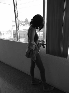 A model wearing a sleek, neutral-toned sleeveless top standing by a sunlit window with natural shadows.