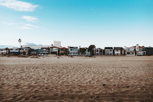 A row of colorful beach houses sits along a sandy coastline under a bright blue sky. The houses vary in architectural styles and colors, creating a picturesque seaside neighborhood. There are palm trees scattered among the houses, hinting at a warm, coastal environment.