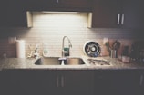 A kitchen countertop with a stainless steel sink and a modern faucet. The countertop has various items including a roll of paper towels, dish soap, a drying rack with utensils, and a few containers. The backsplash is made of light-colored subway tiles, and there are dark cabinets above and below the counter.