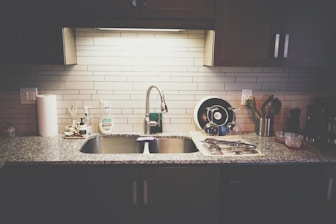 A kitchen countertop with a stainless steel sink and a modern faucet. The countertop has various items including a roll of paper towels, dish soap, a drying rack with utensils, and a few containers. The backsplash is made of light-colored subway tiles, and there are dark cabinets above and below the counter.