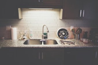 A kitchen countertop with a stainless steel sink and a modern faucet. The countertop has various items including a roll of paper towels, dish soap, a drying rack with utensils, and a few containers. The backsplash is made of light-colored subway tiles, and there are dark cabinets above and below the counter.