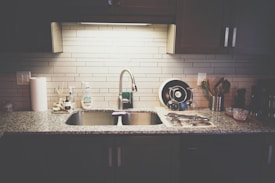 A kitchen countertop with a stainless steel sink and a modern faucet. The countertop has various items including a roll of paper towels, dish soap, a drying rack with utensils, and a few containers. The backsplash is made of light-colored subway tiles, and there are dark cabinets above and below the counter.
