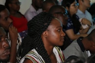 A group of people seated closely together in a dimly lit room, appearing to be attentive and focused. Some individuals are holding devices, indicating engagement with an event or presentation.