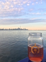 A glass jar filled with an amber-colored beverage is positioned in the foreground against a serene body of water. The distant city skyline is visible under a sky covered in soft clouds, displaying a mix of pastel colors from the setting sun.