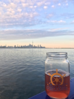 A glass jar filled with an amber-colored beverage is positioned in the foreground against a serene body of water. The distant city skyline is visible under a sky covered in soft clouds, displaying a mix of pastel colors from the setting sun.