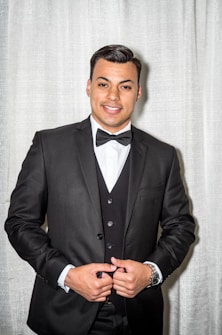 A man is wearing a black tuxedo with a bowtie, standing in front of a light-colored curtain. He has a smile and has his hands adjusting the jacket.