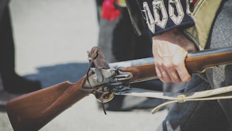 A person wearing historical clothing holds a vintage musket with a wooden stock and metal barrel. The details of the musket's mechanism are visibly intricate, highlighting its historical craftsmanship. The attire features decorative elements, suggesting a period costume, possibly from a reenactment event.
