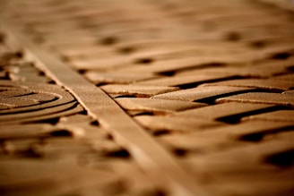 Intricate carvings on a wooden surface are highlighted by soft lighting, creating shadows and depth. The patterns appear geometric and symmetrical, running parallel along the length of the wood.