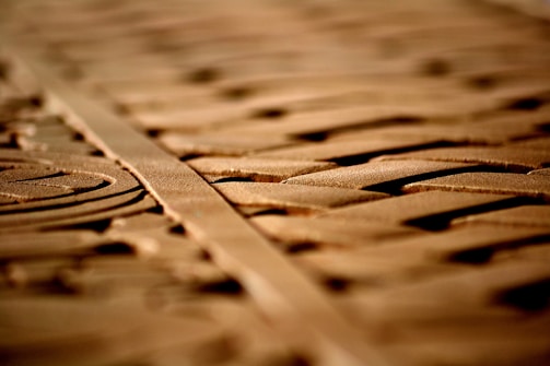 Intricate carvings on a wooden surface are highlighted by soft lighting, creating shadows and depth. The patterns appear geometric and symmetrical, running parallel along the length of the wood.