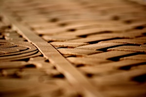 Intricate carvings on a wooden surface are highlighted by soft lighting, creating shadows and depth. The patterns appear geometric and symmetrical, running parallel along the length of the wood.