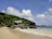 A tranquil beach scene with gentle waves lapping against a sandy shore. A lush, green forest borders the beach, providing a backdrop to a rustic wooden house with a unique roof design. Large boulders are scattered across the sand and into the water, and a few palm trees add to the tropical atmosphere. The sky is bright with fluffy white clouds.