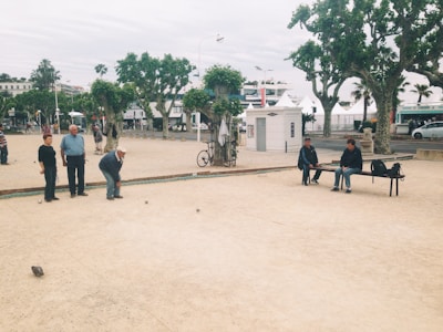 A group of older adults are playing petanque in a public park. Several people are standing by, observing the game, while two individuals are sitting on a bench nearby. The park is lined with large trees and there are bicycles and pedestrians in the background, indicating an urban setting.
