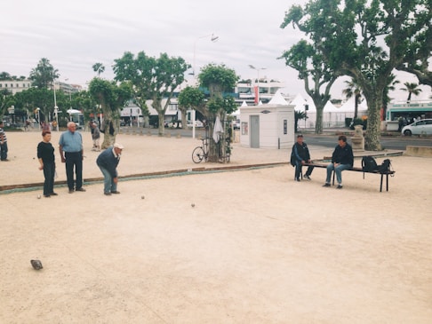 A group of older adults are playing petanque in a public park. Several people are standing by, observing the game, while two individuals are sitting on a bench nearby. The park is lined with large trees and there are bicycles and pedestrians in the background, indicating an urban setting.
