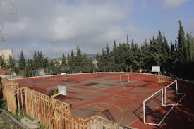 A deserted basketball and soccer court surrounded by a wooden fence and bordered by tall evergreen trees under a partly cloudy sky. The court features multiple hoops, goalposts, and markings for different sports.