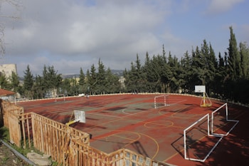 A deserted basketball and soccer court surrounded by a wooden fence and bordered by tall evergreen trees under a partly cloudy sky. The court features multiple hoops, goalposts, and markings for different sports.