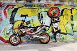A person wearing a motorcycle racing suit and helmet stands with arms crossed beside a sports motorcycle. The background features a vibrant graffiti-covered wall with bold colors and abstract designs.