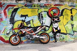 A person wearing a motorcycle racing suit and helmet stands with arms crossed beside a sports motorcycle. The background features a vibrant graffiti-covered wall with bold colors and abstract designs.