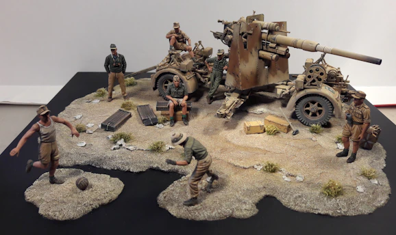 A diorama depicts a World War II scene with several soldiers interacting around a large artillery gun. Some soldiers are seated on a staff car and others stand nearby in various poses. Two soldiers are engaged in a game of soccer on a sandy, rocky terrain. The soldiers wear different uniforms, indicating various roles or nationalities.