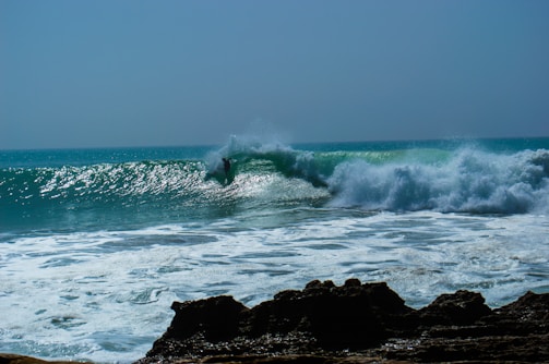 A surfer skillfully rides a large, powerful wave, creating a dramatic splash as the wave curls and crashes. The ocean appears bright and lively under the clear sky, with rocky formations visible in the foreground.