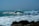 A surfer skillfully rides a large, powerful wave, creating a dramatic splash as the wave curls and crashes. The ocean appears bright and lively under the clear sky, with rocky formations visible in the foreground.