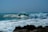 A surfer skillfully rides a large, powerful wave, creating a dramatic splash as the wave curls and crashes. The ocean appears bright and lively under the clear sky, with rocky formations visible in the foreground.