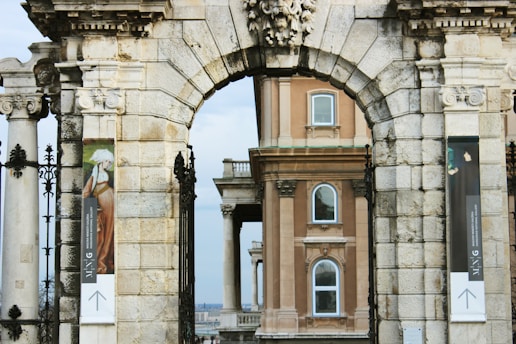 An ornate stone archway frames a view of an elegant, classical building featuring arched windows and elaborate architectural details. There are decorative banners hanging on either side of the archway, depicting art images.
