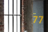 A large window with vertical metal bars is set in an exposed brick wall. Next to the window, there is a gray column with the number 77 painted in large yellow numerals. Above the 77, there is a small yellow label with illegible text.