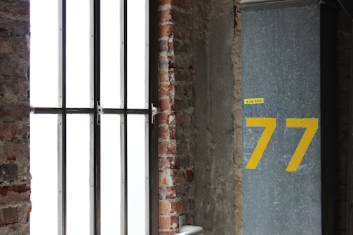 A large window with vertical metal bars is set in an exposed brick wall. Next to the window, there is a gray column with the number 77 painted in large yellow numerals. Above the 77, there is a small yellow label with illegible text.