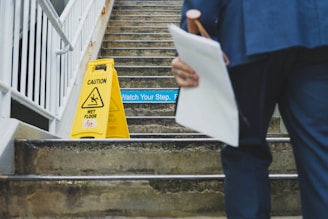 A person holding documents and a pink umbrella is approaching a set of concrete stairs. On the stairs, there is a yellow 'Caution Wet Floor' sign, and a 'Watch Your Step' sign is visible as well. The stairs are metal-railed and seem to be part of a semi-outdoor structure.
