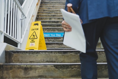 A person holding documents and a pink umbrella is approaching a set of concrete stairs. On the stairs, there is a yellow 'Caution Wet Floor' sign, and a 'Watch Your Step' sign is visible as well. The stairs are metal-railed and seem to be part of a semi-outdoor structure.