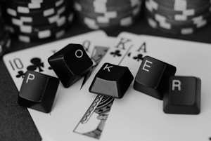 A series of playing cards and poker chips are displayed. The cards, including a ten, jack, queen, king, and ace of clubs, are spread out with black keyboard keys arranged to spell the word 'POKER'. The image is in black and white, giving it a classic, stylish look.