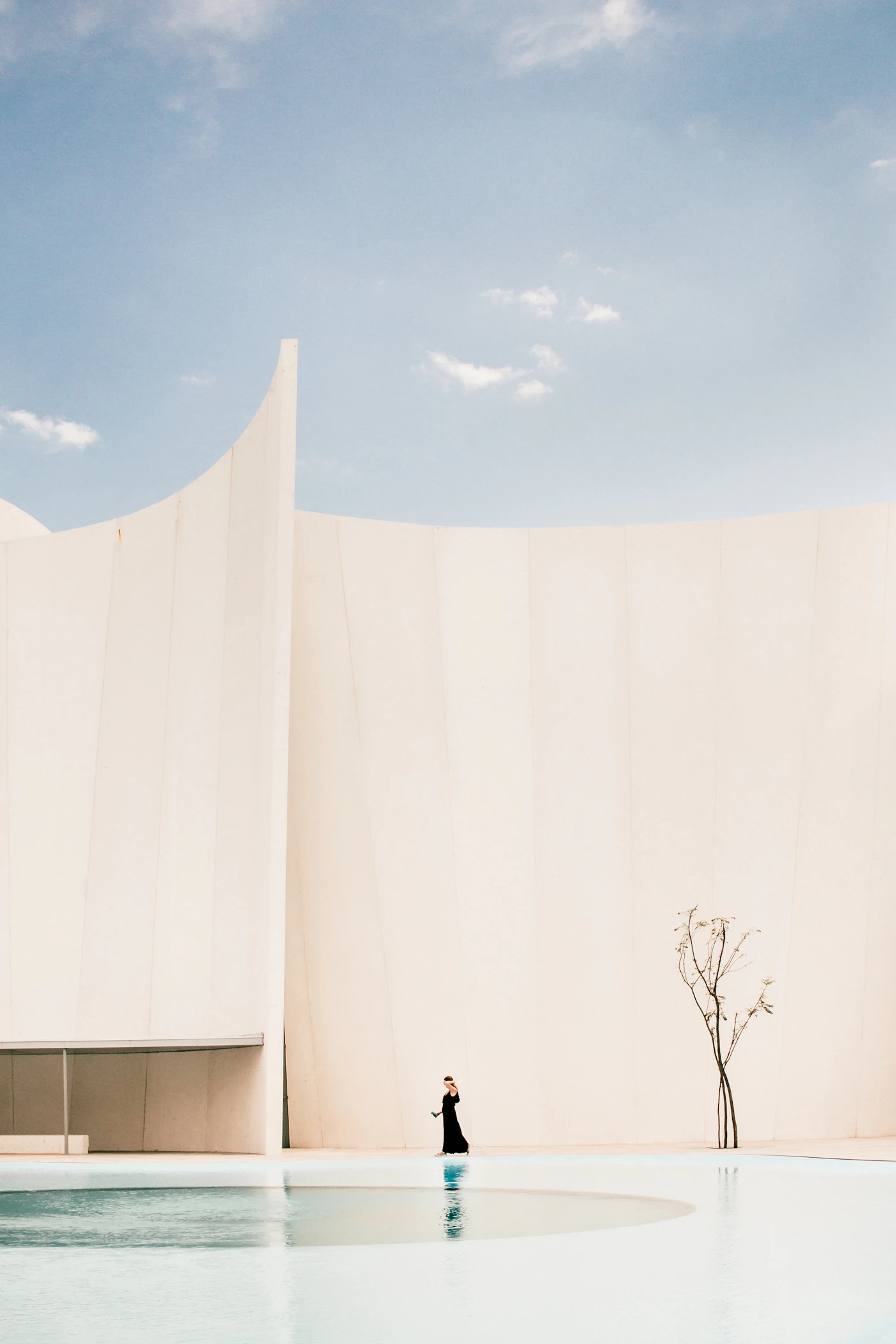 A pale monolithic wall meeting still water, with a lone figure at its edge