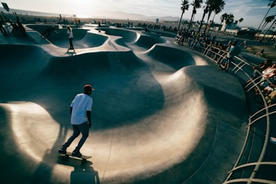 A skatepark scene with multiple skateboarders navigating through curved concrete ramps. The area is surrounded by a crowd of onlookers, and palm trees can be seen in the distance. The lighting suggests it is late afternoon with the sun casting long shadows.