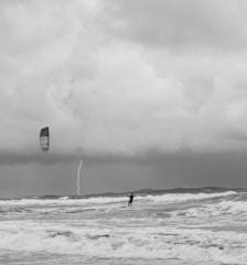 A person is kite surfing on a turbulent ocean with dark, heavy clouds looming overhead. A streak of lightning cuts through the sky in the background, highlighting the dramatic and potentially dangerous weather conditions. The waves appear rough, and the overall scene is monochromatic, adding to the intensity.