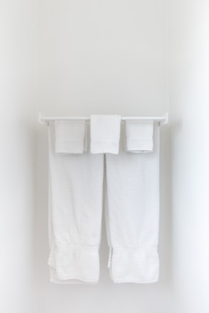 Two neatly folded white towels hang from a clean, white towel rack against a plain, light-colored wall.