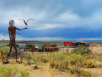A large metal sculpture of a human figure holding a bird is situated in a desert-like landscape. The ground around the sculpture is dry with sparse vegetation. In the background, there is a wooden structure resembling a beachside bar or restaurant with thatched umbrellas. Beyond, there is a vast body of water under a cloudy sky.