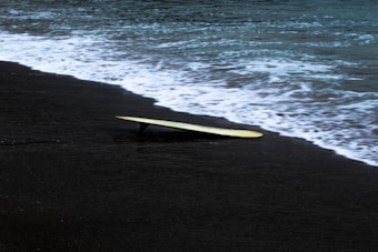 A surfboard lies on a dark sandy beach with waves gently washing ashore, creating a serene coastal scene.
