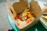 A close-up view of a takeaway box filled with nachos, topped with melted cheese, jalapeños, and diced tomatoes. Next to it, part of a hamburger with lettuce and sauce is visible, set on a plastic tray. The scene is situated on a green table, likely outside.