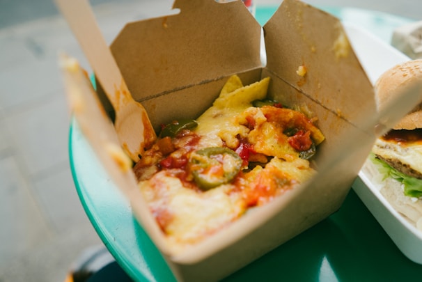 A close-up view of a takeaway box filled with nachos, topped with melted cheese, jalapeños, and diced tomatoes. Next to it, part of a hamburger with lettuce and sauce is visible, set on a plastic tray. The scene is situated on a green table, likely outside.